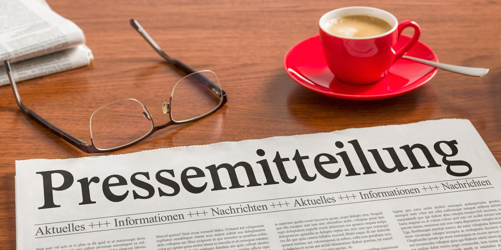Eine Zeitung auf einem Tisch mit dem Titel Pressemitteilung, daneben eine Brille und eine gefüllte rote Expressotasse